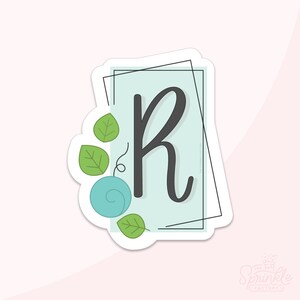 May include: A sticker featuring the letter "R" in a stylish, dark gray script. The letter is set against a light blue rectangle with a black frame. Green leaves and a blue swirl accent the design. The Sprinkle Factory logo is in the bottom right corner.