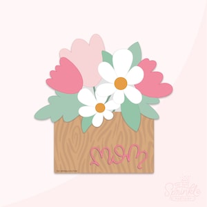 Peut inclure: Une boîte en bois avec un motif de grain de bois et le mot "Maman" écrit en rose. La boîte est remplie de fleurs roses et blanches, dont des marguerites et des tulipes.
