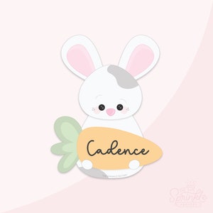 Könnte beinhalten: Ein weißes Kaninchen mit rosa Ohren hält eine Karotte, auf der der Name Cadence steht. Das Kaninchen hat einen grauen Fleck auf dem Rücken. Die Karotte ist orange mit grünen Blättern.