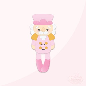 Op de afbeelding: Een roze koekjesvorm in de vorm van een notenkraker met een hoed, uniform en lange benen. De notenkraker heeft een witte snor en wenkbrauwen, met gouden epauletten en twee gouden knopen. Het logo van The Sprinkle Factory staat rechtsonder.