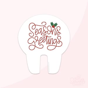 May include: A white cake topper with the words "Seasons Greetings" in red script. The topper has a holly sprig with red berries and a unique shape with two bottom prongs for insertion into a cake. Festive and decorative.