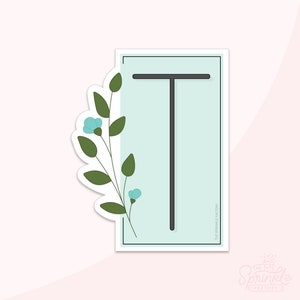 May include: A sticker with a light blue background and a black outline. The letter "T" is in black in the center of the sticker. A green stem with light blue flowers is on the left side of the sticker. The text "THE SPRINKLE FACTORY" is on the right side of the sticker.