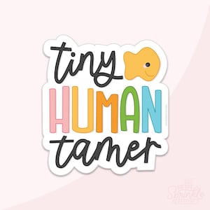 May include: A die-cut sticker featuring the text tiny HUMAN tamer in a mix of black cursive and colorful block lettering. The word tiny and tamer are in black script, while HUMAN is in pink, yellow, orange, green, and blue. A small orange fish icon is included.