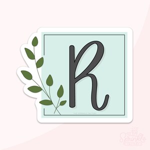 May include: A sticker with a light blue square and a black letter R in a cursive font. A green leafy branch is to the left of the square. The text "THE SPRINKLE FACTORY" is at the bottom of the square.