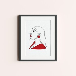 Peut inclure: Une impression encadrée noire d'un dessin au trait d'un visage de profil. La personne porte une chemise rouge et une grosse boucle d'oreille rouge.