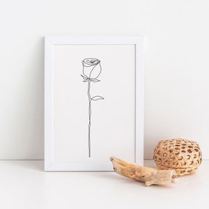 May include: A white frame containing a black line drawing of a single rose. The frame is on a white surface with a woven basket and a piece of driftwood.