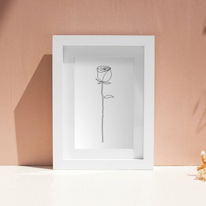 May include: A white frame containing a simple line drawing of a rose. The frame is on a white surface with dried flowers in the background.
