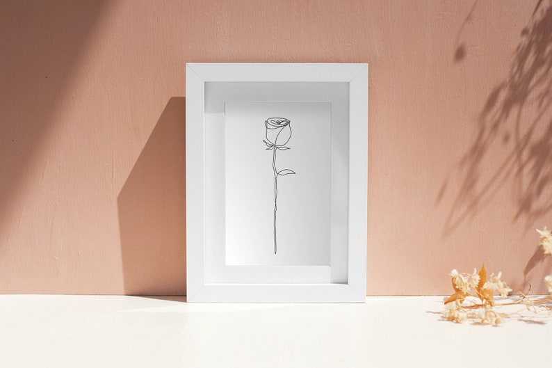 May include: A white frame containing a simple line drawing of a rose. The frame is on a white surface with dried flowers in the background.