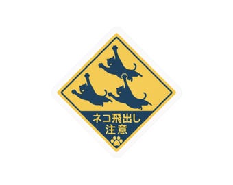 May include: A diamond-shaped yellow and dark blue sticker featuring three dark blue silhouettes of jumping cats. The bottom section contains the Japanese text "ネコ飛出し注意" and a small dark blue paw print icon at the very bottom point.