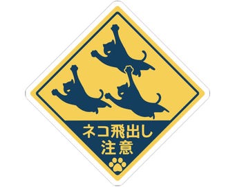 May include: A diamond-shaped yellow and dark blue sticker featuring three dark blue silhouettes of cats leaping in the air. The bottom section contains the Japanese text "ネコ飛出し注意" in yellow, accompanied by a small dark blue paw print icon.