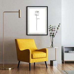 May include: A minimalist line drawing of a single rose in a black frame, hanging on a white wall above a yellow armchair in a living room setting.