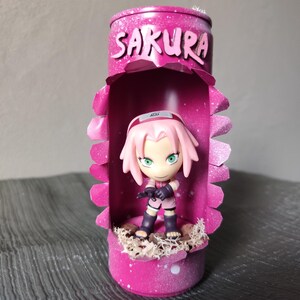 Może przedstawiać: Różowy, dekoracyjny wyświetlacz z figurką Sakura z serii anime Naruto. Wyświetlacz jest wykonany z puszki z recyklingu z napisem "Sakura" białymi literami. Figurka ma na sobie różowy strój.