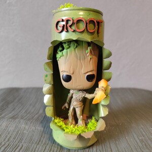 Può includere: Una figura di Groot verde e marrone all'interno di un contenitore personalizzato. Il contenitore ha la forma di una lattina con la scritta "GROOT" in lettere marroni. La figura tiene in mano un oggetto arancione ed è circondata da muschio verde.