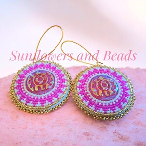 May include: A pair of round earrings with a pink and white beaded design. The earrings have a gold-colored metal frame and a gold-colored charm in the center. The earrings are on a pink surface. The text "Sunflowers and Beads" is in the image.