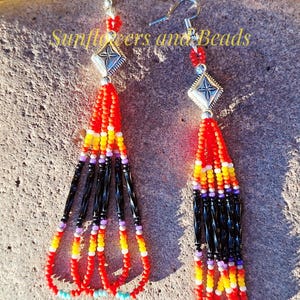 May include: A pair of beaded earrings with a diamond-shaped silver accent. The earrings feature a vibrant color scheme of red, yellow, black, and purple beads. The text "Sunflowers and Beads" is visible in the image.