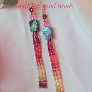 May include: Dangling earrings with gold-colored hooks. Each earring features a rectangular abalone shell bead, pink crystal accents, and beaded strands in shades of pink, red, and gold. The text "Sunflowers and Beads" is visible.