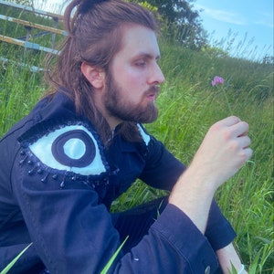 Puede incluir: Una persona lleva una chaqueta vaquera azul oscuro con un gran diseño de ojo en el hombro. La chaqueta tiene lentejuelas negras y detalles blancos. La persona está sentada en un campo de hierba verde y está mirando una pequeña flor morada.