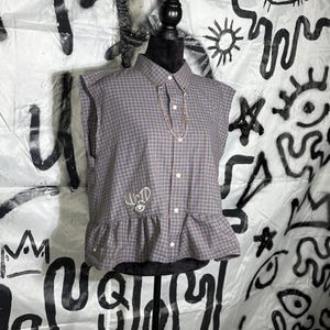 May include: A sleeveless, button-down shirt in a brown and blue plaid pattern. The shirt has a ruffled hem and the word "LUCID" embroidered on the front. A silver necklace is draped on the shirt.