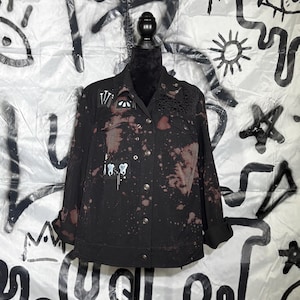Op de afbeelding: Zwarte spijkerjas met bruine, bleekmiddelachtige spetters en witte verfaccenten. De jas heeft een knoopsluiting, een kraag en een klein kanten detail op de rechterschouder. De achtergrond is een wit laken met zwarte graffiti-kunst.