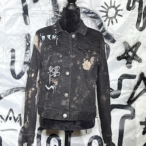 Op de afbeelding: Zwarte spijkerjas met verfspetters, kanten details en het woord "LUCID" in het wit. De jas heeft zilveren knopen en wordt op een mannequin gepresenteerd. De achtergrond is wit met zwarte graffiti-achtige ontwerpen.