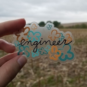 May include: A clear sticker with the word "engineer" in black script, surrounded by orange and teal flower shapes. The sticker is held up against a blurred outdoor background of a field and cloudy sky.
