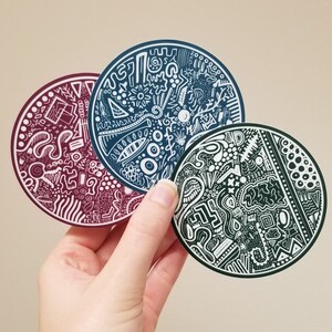 May include: Three circular stickers with intricate white line art designs on a burgundy, navy blue, and dark green background. The stickers are held by a hand against a neutral background. The designs feature abstract shapes and patterns.
