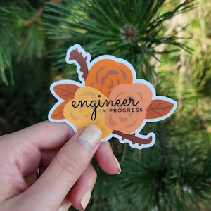 May include: A sticker featuring a floral design in shades of orange, yellow, and brown. The sticker has the words "engineer in progress" in black script. The sticker is held against a backdrop of green pine needles.