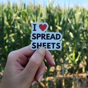 May include: A white sticker with the text "I ❤️ SPREAD SHEETS" in black, with a red heart replacing the word "love". The sticker is held up against a blurred background of green plants.