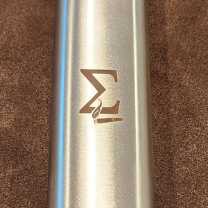 May include: A brushed metal cylindrical container with a logo etched on the front. The logo is a stylized "M" with a flame-like design below it. The container has a smooth, reflective surface.
