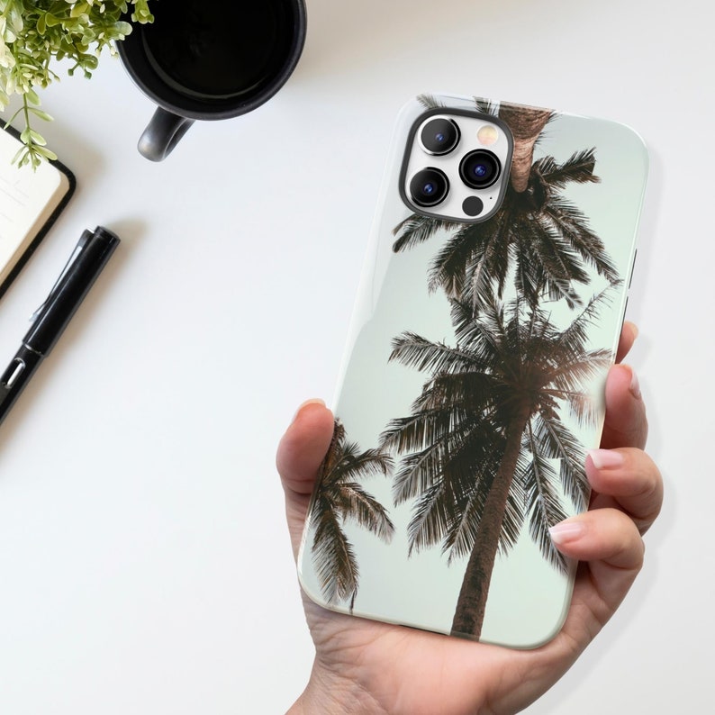 May include: A white phone case with a tropical palm tree design. The case has a camera lens cutout and a white background.