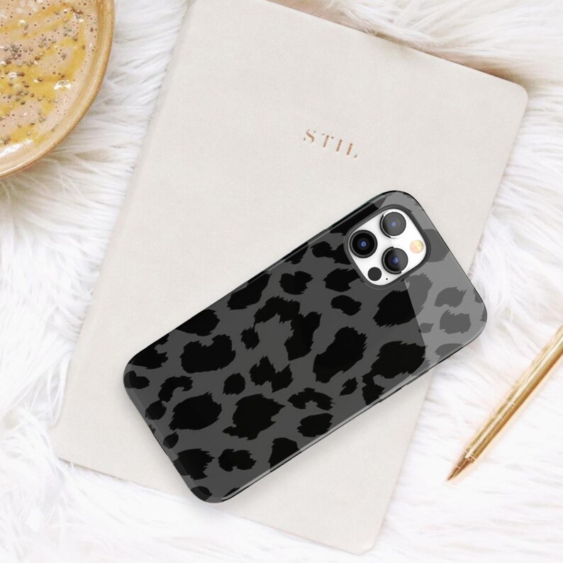 May include: A black and gray leopard print phone case rests on a cream-colored notebook. The phone case has a glossy finish and features a pattern of dark gray spots on a lighter gray background. A gold pen is visible.