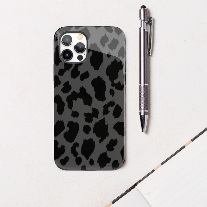 May include: A smartphone case with a gray leopard print design. The case is glossy and has a black camera lens area. A silver pen rests next to the phone case on a white surface.