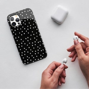 May include: A black phone case with white polka dots. The case is designed for an iPhone with a triple camera lens. The case is shown next to a white wireless charging case and two white wireless earbuds.