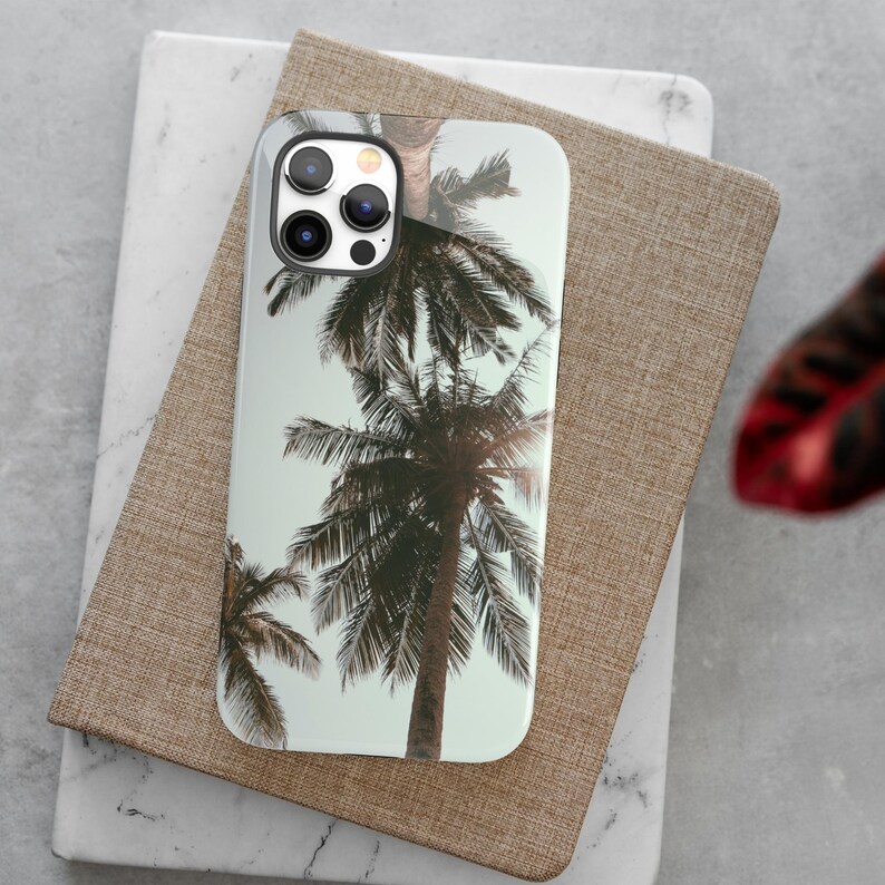 May include: A white phone case with a tropical palm tree design. The case has a camera lens cutout and a black border.