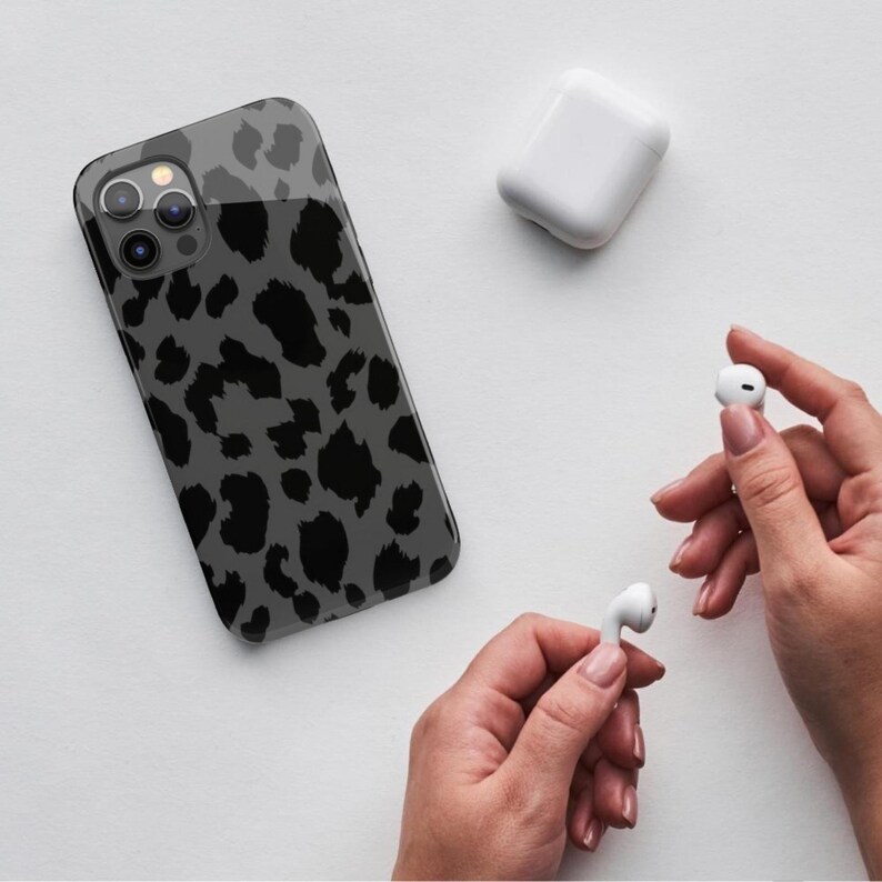 May include: A smartphone case with a gray leopard print design. The case is next to a white charging case and a pair of white wireless earbuds. The background is white.