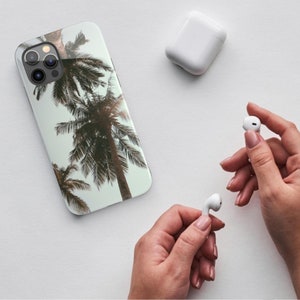 May include: A white iPhone case with a palm tree design. The case has a clear background with brown palm trees and a white outline. The case is shown on a white surface with two white wireless earbuds.