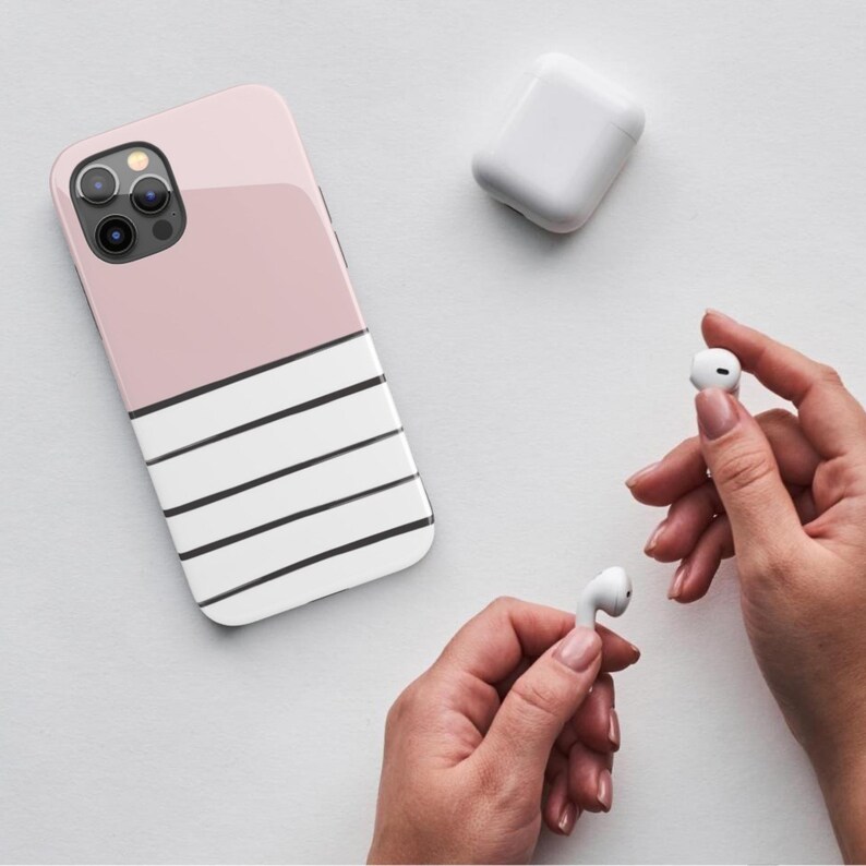 May include: A pink and white striped phone case with a black camera lens. The case is on a white surface with two white wireless earbuds.