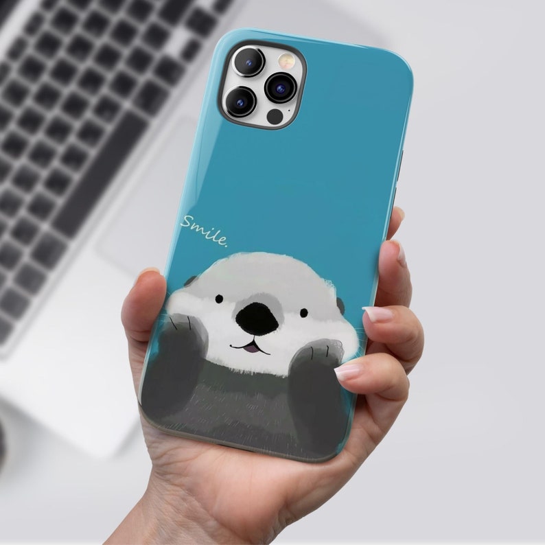 May include: A blue phone case with a cartoon sea otter peeking out from the bottom. The case has a white inscription that says "Smile".