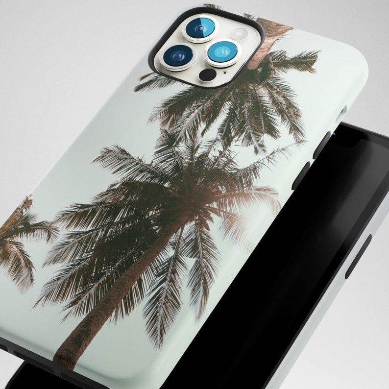 May include: A white iPhone case with a tropical palm tree design. The case has a camera lens cutout with three black camera lenses.