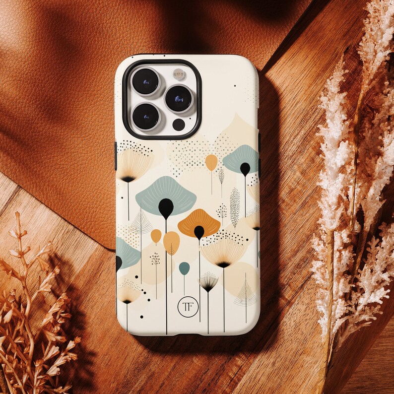 K&ouml;nnte beinhalten: Eine wei&szlig;e iPhone-H&uuml;lle mit einem geometrischen Muster aus abstrakten Blumen in Gelb-, Braun- und Blaut&ouml;nen. Die H&uuml;lle hat ein klares Kameraobjektiv und den Text "TF" in der Mitte.