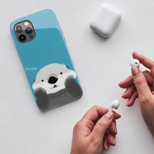May include: A blue phone case with a cartoon sea otter on it. The otter is smiling and has its paws up to its face. The text "Smile." is printed on the case.