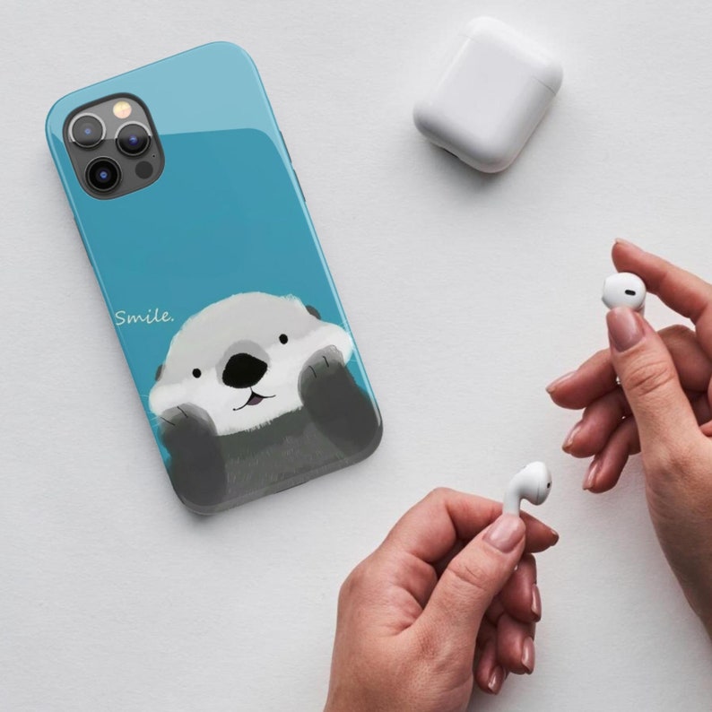 May include: A blue phone case with a cartoon sea otter on it. The otter is smiling and has its paws up to its face. The text "Smile." is printed on the case.