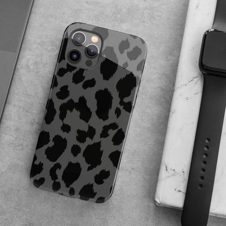 May include: A smartphone case with a gray leopard print design. The case is black and gray with a glossy finish. The phone case is next to a black smartwatch and a laptop.