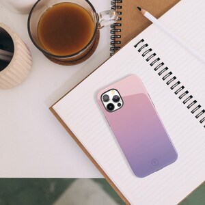 May include: A pink and purple gradient phone case with a camera lens cutout and a small circle logo. The case is lying on a notebook with lined paper.