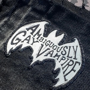 Op de afbeelding: Een witte geborduurde patch in de vorm van een vleermuis, met de zwarte tekst "AMBIGUOUSLY GAY VAMPIRE" op de stof gestikt. De patch is bevestigd op een stuk zwarte spijkerstof met een gerafelde rand aan de bovenkant van het materiaal.