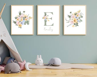 May include: Three framed floral art prints with a light wood frame. The prints feature watercolor bouquets in pastel colors and a large green letter "E" with the name "Emily". The prints are displayed above a wooden floor.