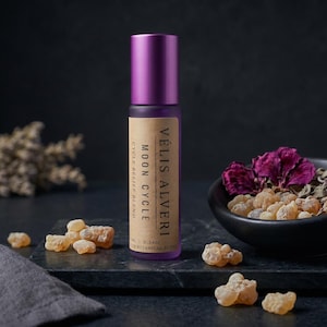 May include: A purple essential oil roller bottle with a matching cap, labeled "Vélis Alveri Moon Cycle Cycle Relief Blend." The image includes a black bowl with dried flowers and herbs, and scattered amber-colored resin pieces.