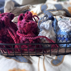 May include: A black metal basket holds several crocheted drawstring bags. The bags are in shades of red, white, and blue. The basket sits on a patterned fabric, creating a cozy, handmade aesthetic.