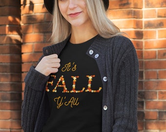 May include: A woman wearing a black t-shirt with the text "It's Fall Y'all" in gold letters with a fall leaf pattern. She is wearing a black cardigan, black pants, and black boots. She is standing in front of a brick wall.