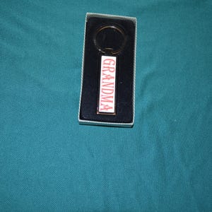 May include: A rectangular keychain with the word "GRANDMA" in red capital letters on a white background. The keychain is in a black box with a silver border, set against a teal fabric.